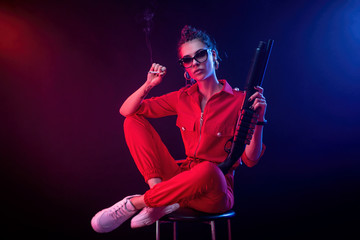 Fototapeta premium jasna brunetka w czerwonym kombinezonie ze strzelbą na ciemnym tle w świetle neonów