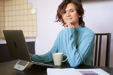 Portrait of young girl looks at camera, leans on chin, sitting in quarantine during pandemic. Beautiful brunette Bob hair wears blue sweater. coffee mug, paper, Notepad, free space for inserting text