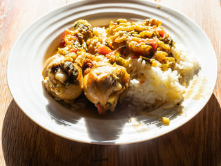 portion of Chakhokhbili (traditional georgian dish from stewed chicken, tomato, eggplant and fresh herbs) with boiled rice on white plate on wooden table in home kitchen backlighted by sun