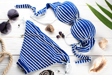 Separate blue and white striped swimsuit on a white wooden background of boards, next to accessories, shells, glasses, earrings