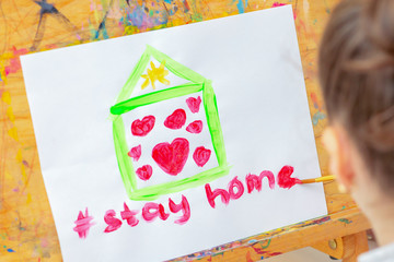 Hand of child drawing green house with red hearts and words Stay Home with brush on sheet of paper during quarantine at home. Child drawing during education.