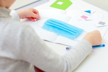 Hands of child holding a medical mask over copybook during studying at home. Home learning concept.