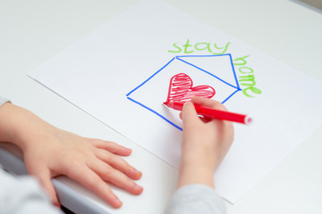 Hands of child drawing red heart inside house with words Stay Home on white sheet of paper. Stay Home concept.