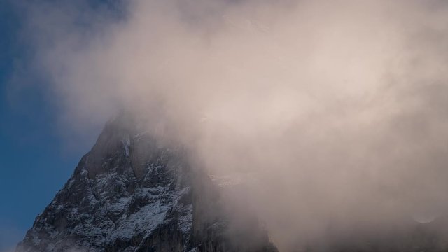 Time lapse of misty clouds rolling over the snow covered mountain peak of the Eiger North Face with blue sky in the background in Grindelwald, Switzerland