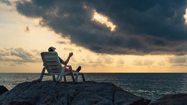 A man with a glass in his hand sits in a lounger on the rocs by the sea against the background of dawn on a summer morning. 4K time-lapse