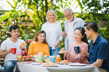 Fototapeta na wymiar Happy big family have a lunch at outdoor in green garden. Lunch or tea time on picnic table in summer. Old woman make a bread with Fruit jam to give husband. Big family outdoor lunch concept.
