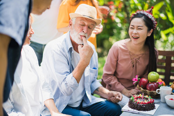 Happy big family smiling have a lunch at outdoor in green garden. Grand father's birthday party on picnic table in summer. Old man test the cake by use finger. Big family outdoor lunch party concept.