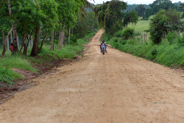 A man on a bike carrying a passenger and travelling on a dirt road in the bush