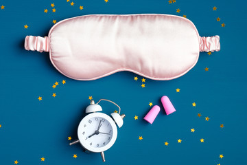 Sleep eye mask with alarm clock and ear plugs on dark blue background. Good night, sleep hygiene,...