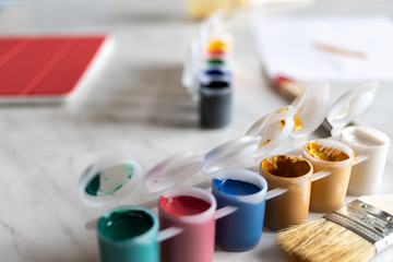 paints of different colors on a light table, a brush next to it. on the table is a tablet with a red cover. there is a drawing sheet in the distance. natural light and shadows