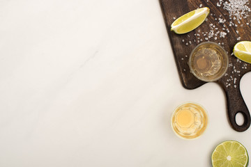 top view of golden tequila with lime, salt on wooden cutting board on white marble surface