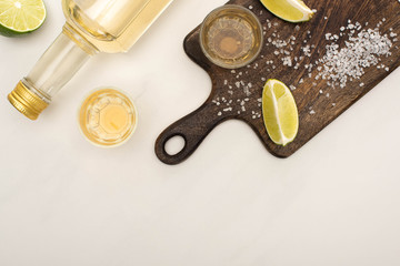 top view of golden tequila with lime, salt on wooden cutting board on white marble surface