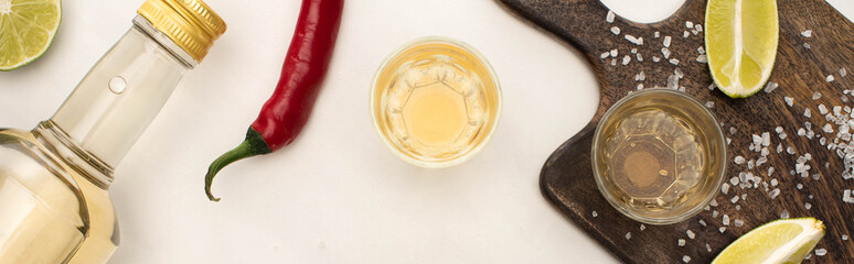 top view of golden tequila with lime, chili pepper, salt on wooden cutting board on white marble surface, panoramic shot
