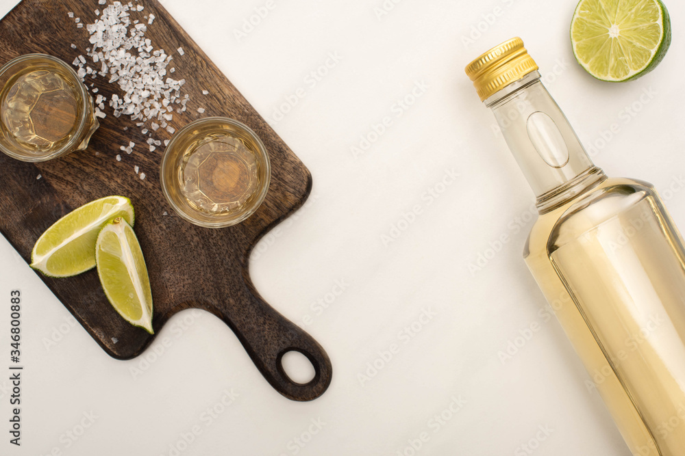 Poster top view of golden tequila with lime, salt on wooden cutting board on white marble surface