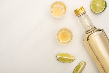top view of golden tequila in bottle and shot glasses with lime on white marble surface