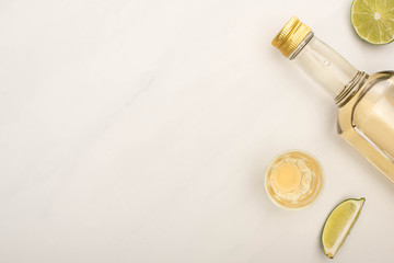 top view of golden tequila in bottle and shot glass with lime on white marble surface