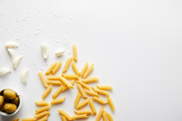 Top view of pasta, garlic, bowl with olives and sea salt on white background
