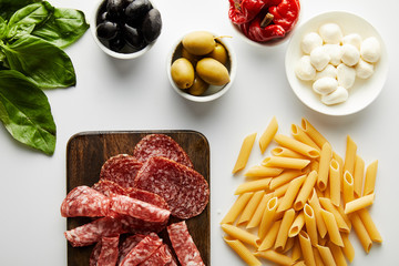 Top view of meat platter, pasta, basil leaves and bowls with ingredients on white background