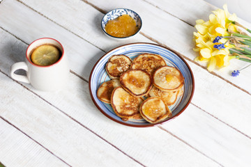 Fritters in blue plate, honey with spoon, cup of coffee with lemon, daffodile flowers bunch on white wooden background