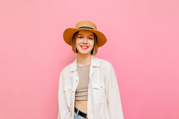 Happy tourist girl in hat and in summer clothes stands on pink background and poses at camera with a smile on her face. Cheerful lady in light light clothing isolated on pastel background. Copy space