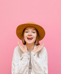 Portrait of a joyful girl in a hat and light summer clothes stands on a pastel pink background and rejoices with palms raised, looks in camera and shouts. Isolated. Vertical