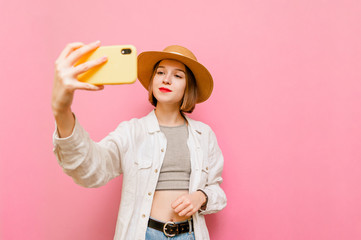 Pretty girl in summer light clothing poses on smartphone camera, makes selfie on pink background. Attractive lady in hat and light clothing takes selfie on smartphone. Isolated.