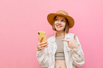 Expressive girl in hat shows finger on smartphone and looks away at copy space with positive face on pink background. Tourist girl with smartphone in hands and with shocked face isolated on pink