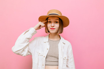 Pretty girl with a serious face stands on a pink background and points finger at the temple,...
