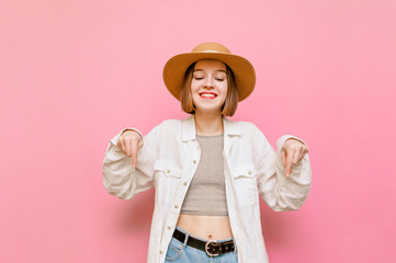 Smiling lady in hat and light clothing isolated on pink background, shows and looks down at copy space. Happy girl tourist points hands down while standing against pink background.