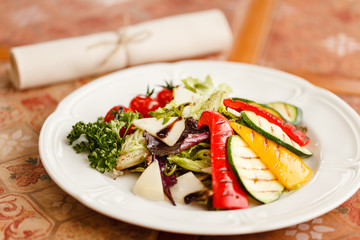 grilled vegetables with cheese and herbs, seasoned with balsamic vinegar. served on a white round plate on a wooden table top decorated with ceramic tiles