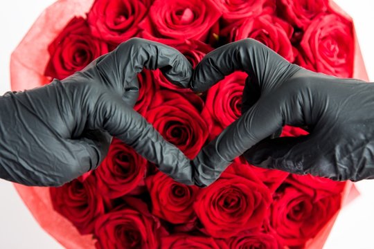 Hands in medical gloves making sign Heart by fingers against red roses, Selective focus. Contactless flower delivery during isolation during coronavirus pandemic with love