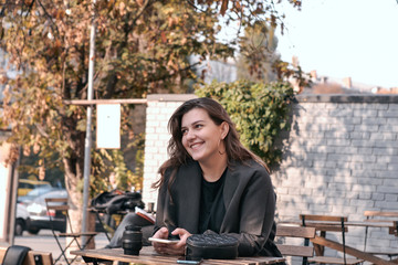 young woman  sits at a table on a summer terrace of a cafe. she is smiling. she holding smart phone in her hands