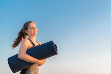 A young millennial smiling girl in sportswear is standing on a blue sky background, holding a rolled mat in her hands