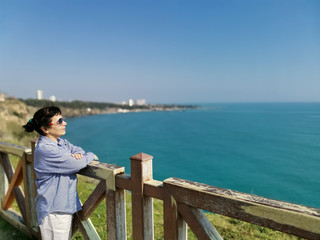 A middle-aged woman in a shirt and sunglasses stands on a high bank above the sea and looking at great distances.