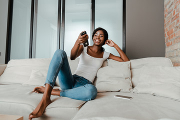 Obraz premium African american woman smiling with remote controller near smartphone on sofa in living room