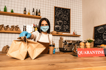 Cafe owner in medical mask with disposable cup of coffee showing paper bags near table with card...