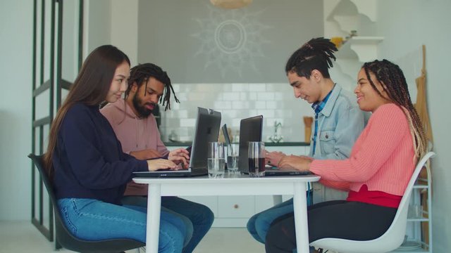 Group of diverse multiracial college students gathered at table in domestic kitchen, learning together and communicating, networking with digital devices while preparing for university exams