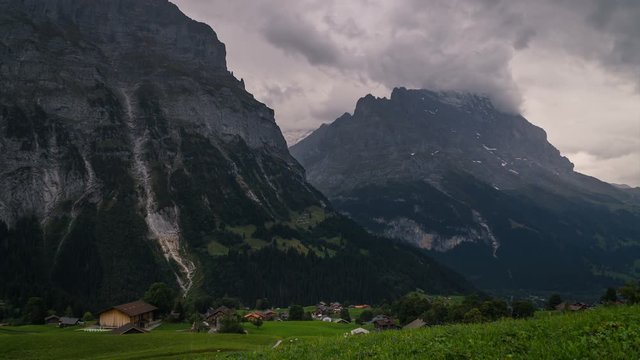 Time lapse of dark storm clouds moving over the Eiger mountain ranges in the Swiss Alps of Grindelwald Switzerland. The green farmland valley with alpine houses is dwarfed by the tall mountain peaks.
