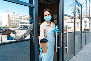 Cafe owner in medical mask near door showing disposable cup of coffee and looking at camera on street