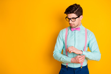 Photo of handsome cool clothes young guy boyfriend look empty space seriously buttoning shirt sleeve wear specs teal shirt pink suspenders bow tie isolated yellow color background