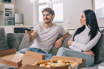 emotional couple having pizza and watching tv with remote controller during self isolation at home