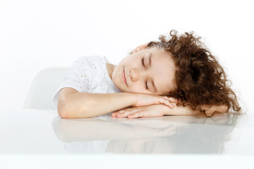 Obraz premium Tired little curly girl sleeping on the desk, over white background.