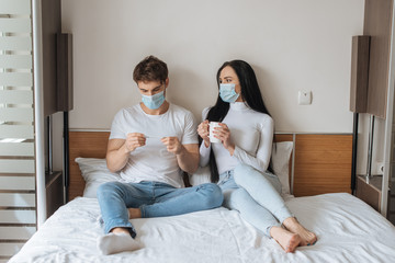 Worried couple in medical masks holding cup and thermometer in bedroom during self isolation