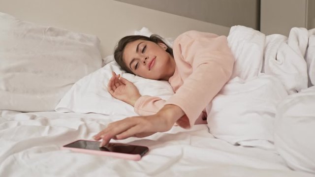 A young sleepy girl is turning off her alarm clock on her mobile phone laying in the bed at home