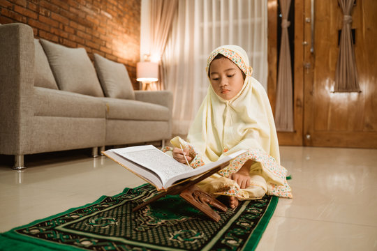 cute asian girl reading quran by herself at home. muslim toddler with hijab