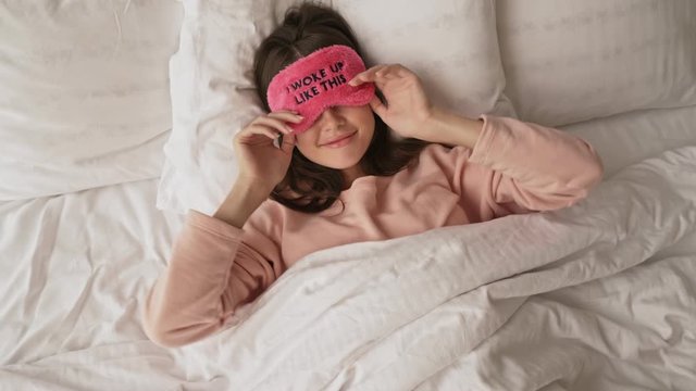 A lovely cheerful girl is taking off her sleeping mask laying in the bed at home