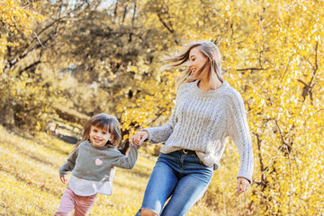 Fototapeta premium Beautiful woman and her little daughter holding hands and running in autumn park. Tiny girl having fun and playing with her young mother outdoors. Happy family relationships of mom and child