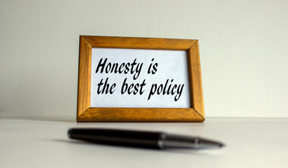 Wooden picture frame with inscription 'honesty is the best policy' on beautiful white fon. Pen on the table.