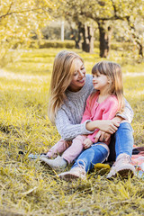 Happy young mother sitting on blanket together with her beautiful daughter and smiling outdoors. Cheerful woman playing with her little girl in autumn park. Girl playing with mom. Family love