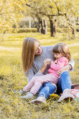 Fototapeta na wymiar Beautiful woman playing with her young daughter and laughing while sitting on blanket in autumn park. Little girl hugging with her young mother and smiling outdoors. Family relationships and love